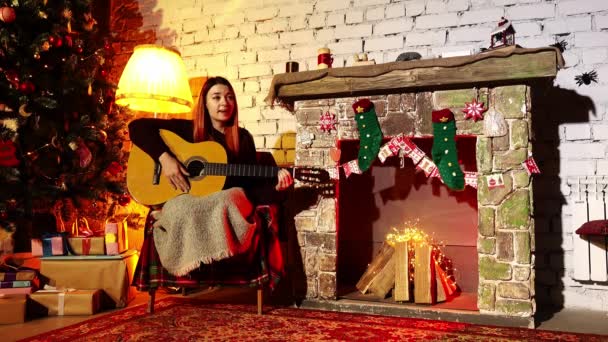 Uma mulher passa férias de inverno tocando guitarra em casa em um ambiente acolhedor com um abeto e lareira — Vídeo de Stock