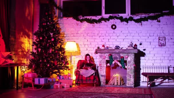 Vrouw ontspant in fauteuil leesboek met kerst in de buurt van open haard en dennenboom — Stockvideo