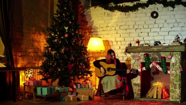 Женщина проводит зимние каникулы играя на гитаре дома в уютной атмосфере с пихтой и камином — стоковое видео