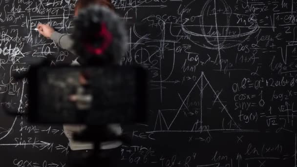 A young teacher gives an online math lesson in front of a smartphone camera standing at chalkboard — Stock Video