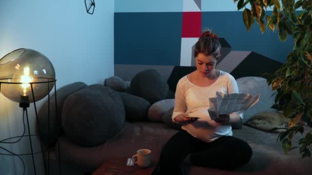 Νεαρή έγκυος καυκάσια γυναίκα κάθεται στον καναπέ στο άνετο σαλόνι και διαβάζοντας χάπια συνταγή — Αρχείο Βίντεο