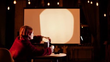 Bayan rahat bir odada koltukta oturuyor ve fotoğraf negatiflerini slayt projektörüne yerleştiriyor.