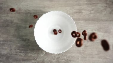 Çikolatalı gevrek halkaları, ağır çekimde tahta bir masanın üzerinde duran beyaz bir kaseye düşüyorlar.