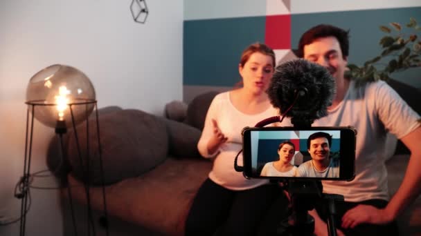 Man och gravid kvinna ger en intervju sittande på en soffa i vardagsrummet framför mobilkameran — Stockvideo