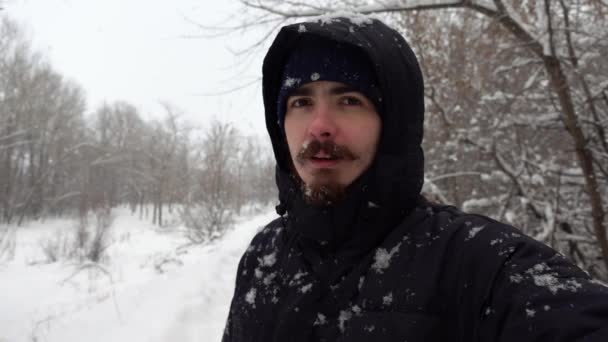 Молодой кавказский мужчина делает селфи стоя в зимнем лесу с падающими снежинками в медленном темпе — стоковое видео