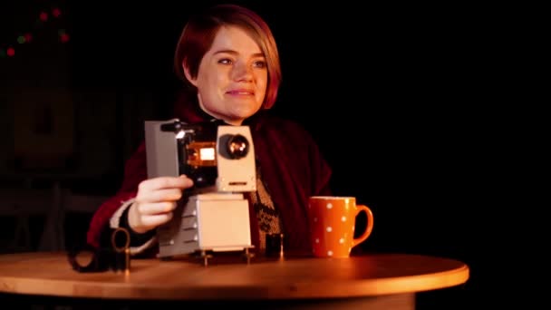 Usmívající se žena sedí u stolu a sleduje skluzavky s pomocí retro projektoru — Stock video