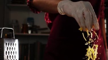 Kırmızı üniformalı ve koruyucu eldivenli bir şef yavaş pişirme sürecinde rendelenmiş peynir ekliyor.