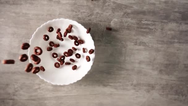 Anéis de cereais de chocolate derramando em placa branca de pé sobre mesa de madeira em câmera lenta — Vídeo de Stock