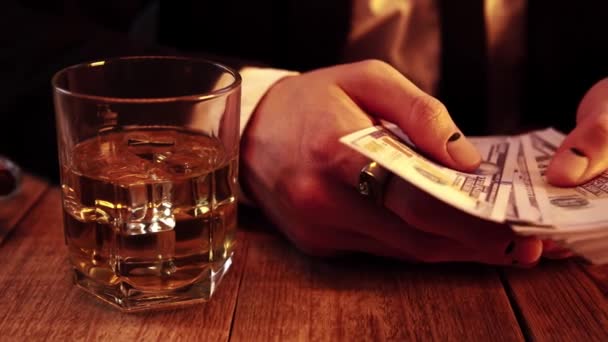 Uomo ricco in costume nero conta un fascio di soldi seduto a un tavolo di legno accanto a un bicchiere di whisky — Video Stock