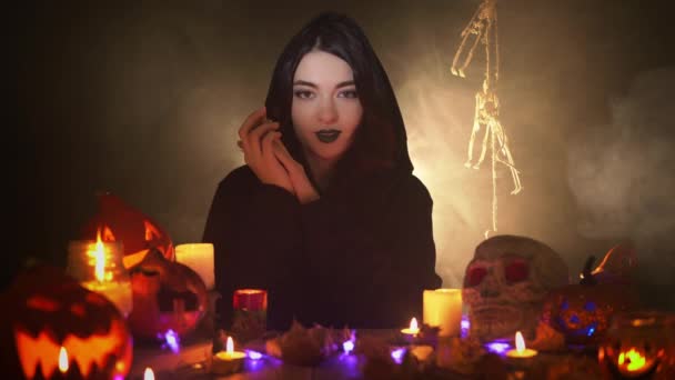 Mystical woman in a dark mantle sits among candles and pumpkins surrounded by smoke — Stock Video