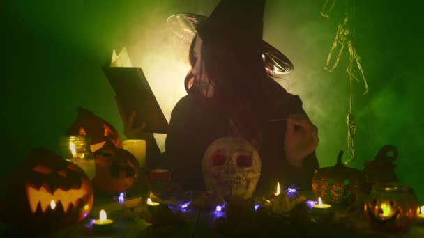 Mladá gotická čarodějnice v tmavém plášti a klobouku vykouzlí nad lebkou mezi halloweenskými dekoracemi — Stock video