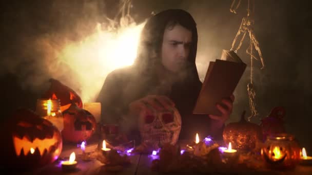 Молодий чарівник практикує чаклунство над черепом, читаючи чарівну книгу на чорному тлі — стокове відео