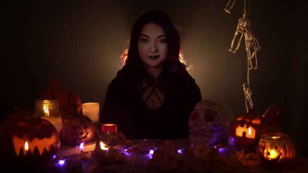 Gothic witch shows thumb up sign sitting among jack olanterns and candles on black background — Stock Video