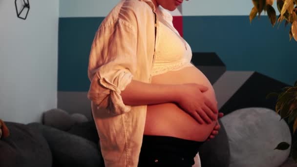 Καυκάσια έγκυος γυναίκα στέκεται σε ένα σαλόνι με τέχνη εσωτερικό και χαϊδεύοντας τρυφερά κοιλιά — Αρχείο Βίντεο