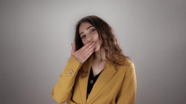 Молодая женщина в желтой куртке с длинными вьющимися волосами отправляет воздушный поцелуй в медлительности на белом фоне — стоковое видео