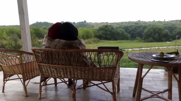 Un couple amoureux est assis sur la terrasse sur banc de canne étreignant et couvrant de couverture — Video