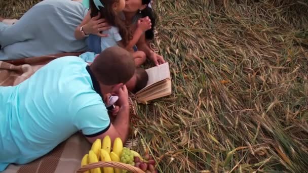 Familie bij de picknick ligt op de grond van een tarweveld met het boek — Stockvideo