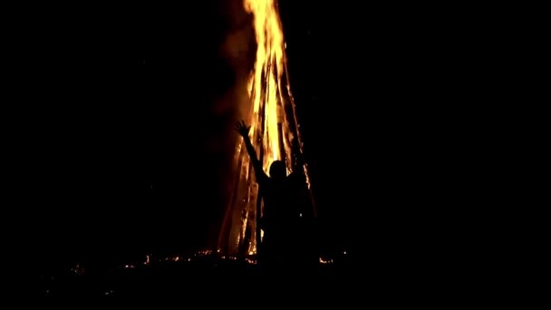 Achteraanzicht van een man die voor een enorm brandend kampvuur staat en met zijn handen zwaait in slow motion — Stockvideo