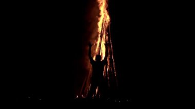 A man standing in the darkness in front of a big bonfire is holding his hands up and praying on it