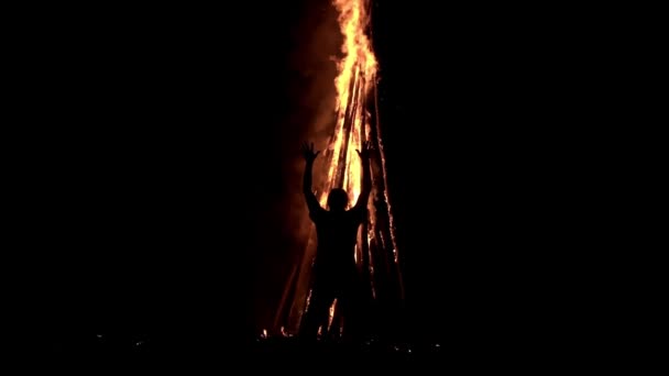 A man standing in the darkness in front of a big bonfire is holding his hands up and praying on it — Stock Video