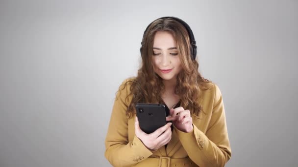 Attraktiv kvinna i hörlurar välja musik i telefon och danser står på en vit bakgrund — Stockvideo