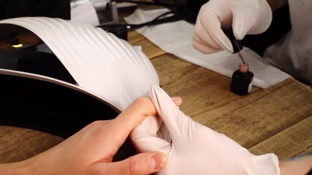 Uma manicure fêmea cobre as unhas masculinas do cliente por gel polonês e diz para colocar as mãos na lâmpada UV — Vídeo de Stock