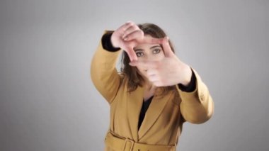 Beyaz bir arka planda duran ve çerçeveyi parmaklarıyla gösteren gülümseyen genç bir kadın portresi