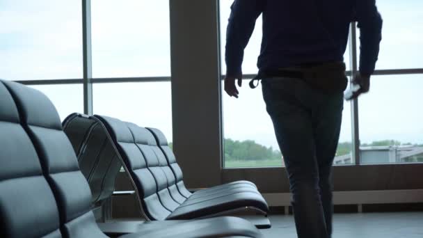 El hombre viene en un vestíbulo del aeropuerto internacional y se sienta en el banco con el teléfono inteligente en la mano — Vídeos de Stock