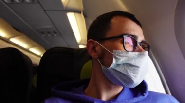 Tıbbi maskeli bir adam uçağın tahtasına oturmuş pencereden izliyor.
