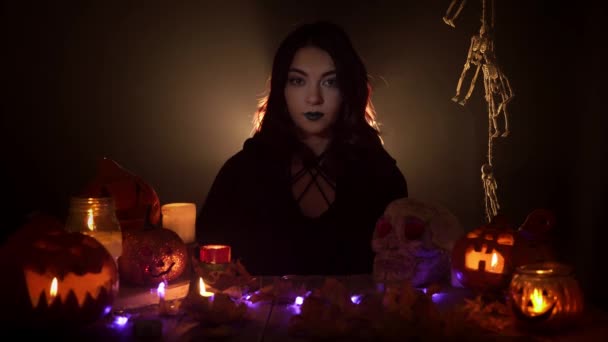 Une jolie sorcière souriante montre un panneau Ok assis parmi les décorations d'Halloween sur le fond noir — Video