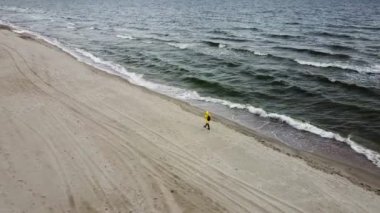 Dişi turist, kumlu sahilde, okyanusun kıyısında yürüyor. Rüzgarlı ve soğuk havada, yukarıdan çekilmiş.