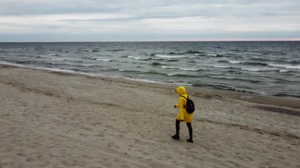 Женщина в желтом пальто с мешком на спине идет по берегу океана в холодный ветреный день — стоковое видео