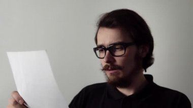 Gözlüklü, ciddi bir genç, kağıda basılmış bir belgeyi okuyor ve üzgün görünüyor.