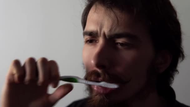 Vista de cerca del hombre cansado barbudo limpiando sus dientes con cepillo y pasta de dientes sobre fondo blanco — Vídeos de Stock