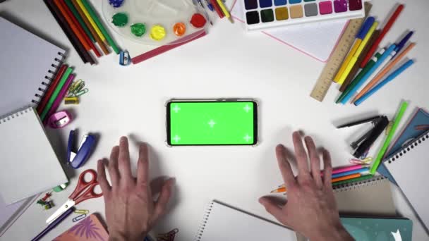 Vedere de sus a mâinilor omului bătând cu degetele pe masă plină de accesorii școlare în fața unui mobil — Videoclip de stoc