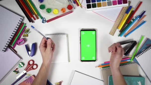 Hombre pasa hacia arriba y se acerca a un teléfono inteligente con pantalla verde que se encuentra en el escritorio blanco entre las herramientas de la escuela — Vídeos de Stock