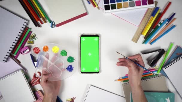 El hombre sostiene la paleta con colores y el pincel sobre el teléfono inteligente con pantalla verde — Vídeos de Stock