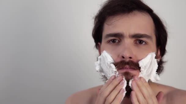 Человек кладет пену на лицо и бреет бороду металлическим кухонным ножом, стоя на белом фоне — стоковое видео