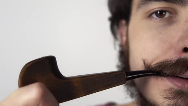 Closeup footage of young man with curly mustache smoking pipe standing on white background — Stock Video