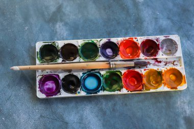 Çeşitli renklerde eski suluboya resimler ve beyaz plastik bir kutuda kirli bir fırça. Seçici odaklanma. Odadaki gri arkaplan