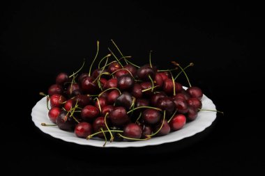 Masadaki beyaz bir tabakta kırmızı kirazlı olgun, sulu siyah arka plan seçici odak noktası görünümü.