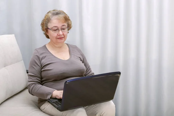 An elderly woman sits in a room on the couch and works at a laptop — Stock Photo, Image