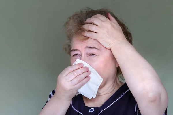 An elderly woman cries, her head hurts, she holds her head with her hand and presses a napkin to her face, — Stock Photo, Image