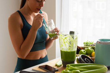 Mutfakta yoga kıyafetiyle yeşil smoothie içen bir kadın telefonu kontrol ederken...