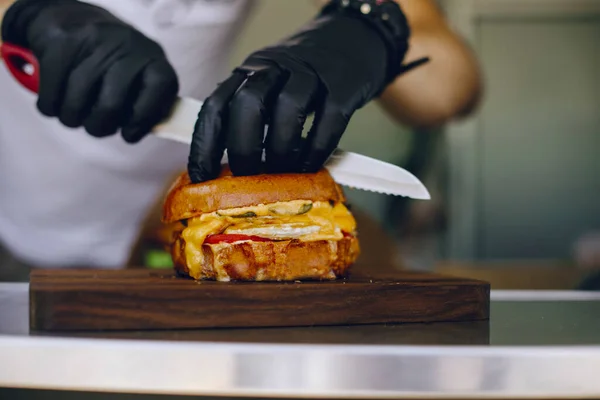 Siyah eldivenli şef bıçakla burger kesiyor.