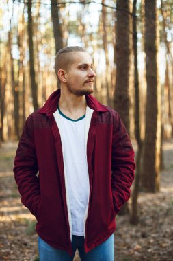 Beyaz tişörtlü ve kırmızı ceketli sakallı adam, orman parkının arka planındaki bulanık arka plan bokeh etkisine güvenle bakıyor.