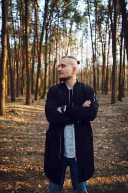 Tarz sahibi sakallı bir adam kolları çaprazlanmış bir şekilde çam ormanında dikiliyor ve uzaklığa bakıyor.