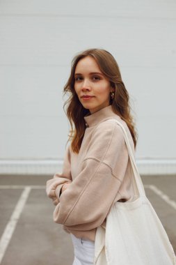 Dalgalı saçlı genç bir kız park yerindeki beyaz duvarın yanında beyaz bir çevre çantasıyla duruyor.