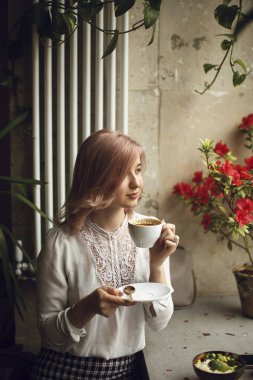 Beyaz bluzlu genç bir kadın rüyasında yan tarafa bakıyor ve kafeteryada kahve ile birlikte beyaz bir fincan tutuyor.