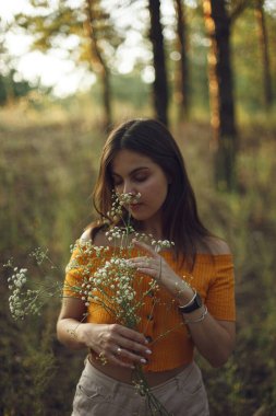 Ormanda gün batımında turuncu tişörtlü genç bir kızın ellerinde kır çiçeği.